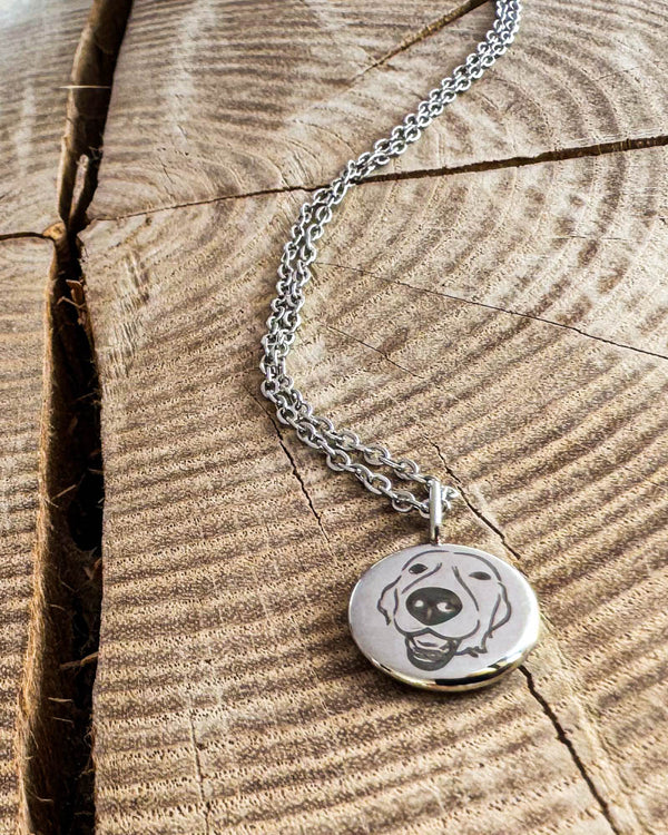 AYA PORTRAIT necklace in silver-coloured steel with a drawing of a happy dog engraved on a small pendant, on light wood with a crack