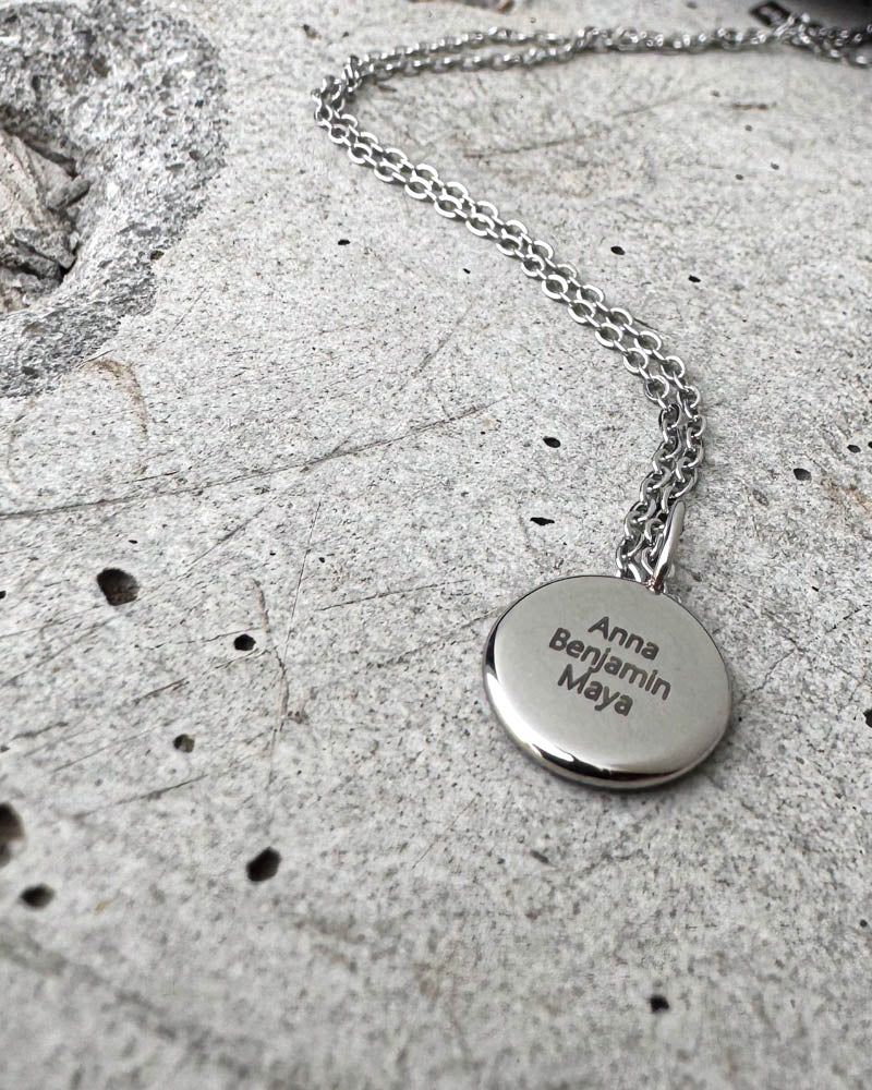 AYA QUOTE name necklace in silver-coloured steel with the names "Anna Benjamin Maya" engraved on a small round pendant, on concrete