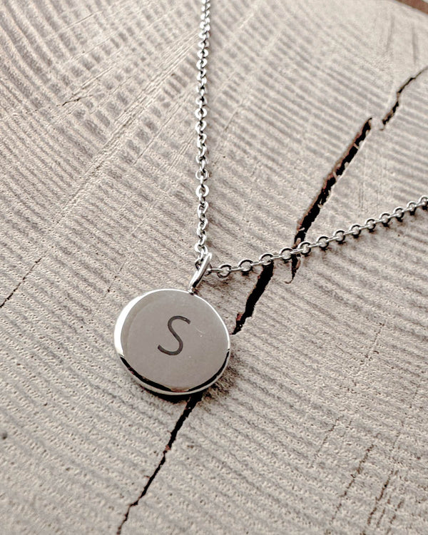 AYA LETTER necklace in silver-coloured steel with the letter "S" engraved on a small round pendant, on light wood with a crack