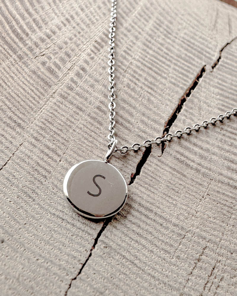 AYA LETTER necklace in silver-coloured steel with the letter "S" engraved on a small round pendant, on light wood with a crack
