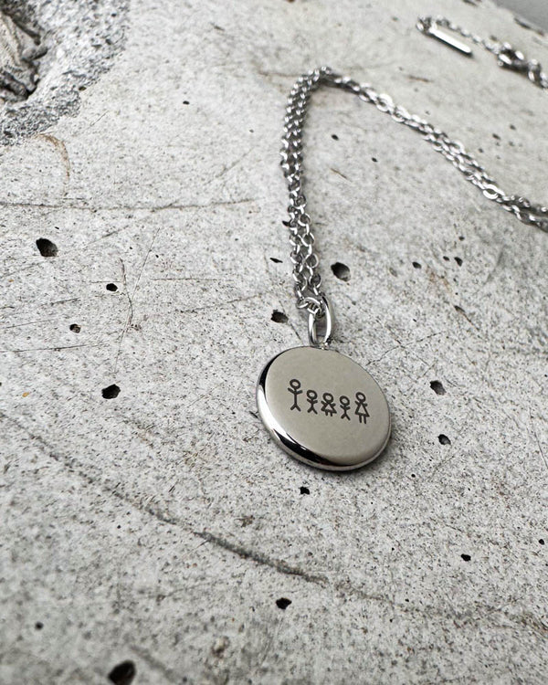 AYA STICK necklace in silver-coloured steel with a matchstick family of five engraved on a small round pendant, lying on concrete