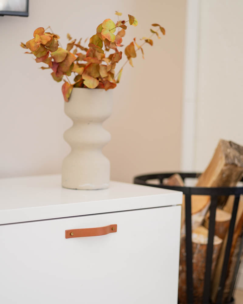 BAND leather drawer pull in cognac on a white chest of drawers with beige vase of autumn branches and log basket beside it