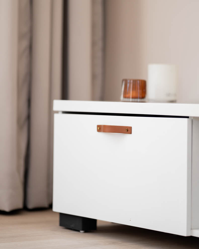 BAND leather drawer pull in cognac with brass screws on a white TV unit drawer, scented candle and light curtains behind