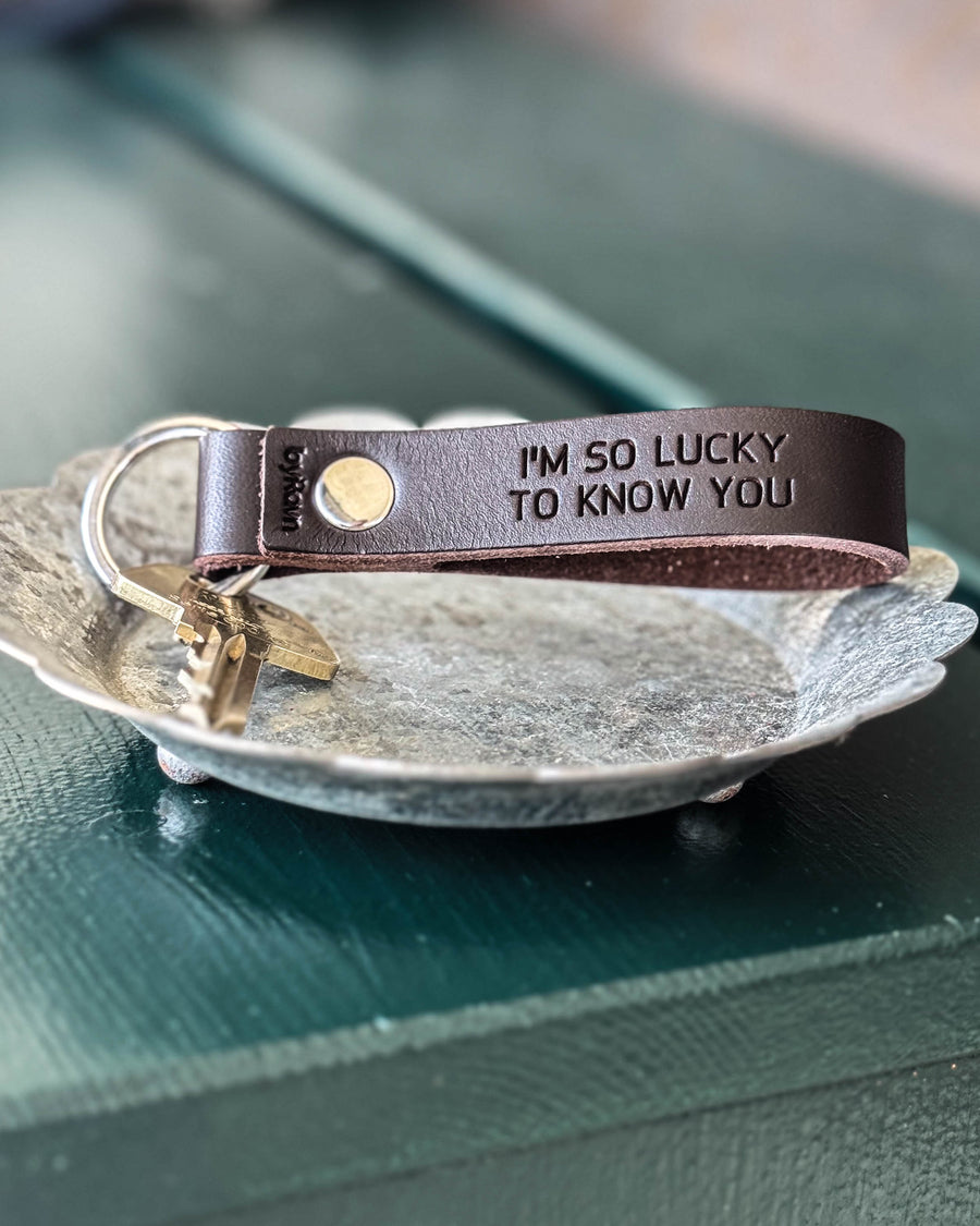 BOND leather keyring in dark brown with the engraving "I'm so lucky to know you" and key in a zinc dish on a green table