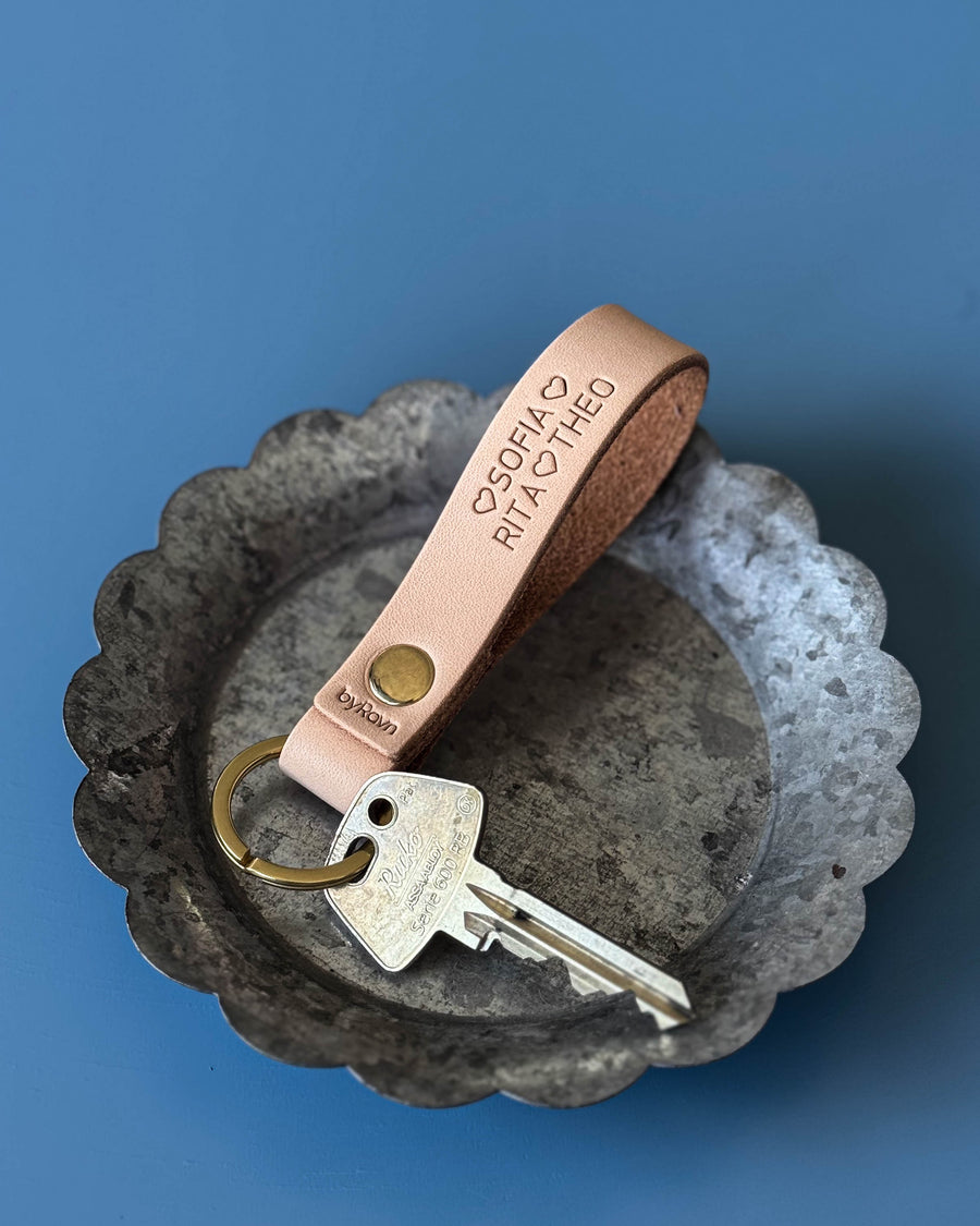 BOND leather keyring in natural with the names "Sofia, Rita, Theo" ♡ engraved, in a zinc dish with key on a blue background