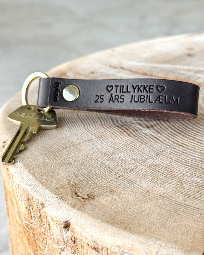 BOND leather keyring in dark brown with the engraving "Tillykke 25 års jubilæum" ♡ and brass rivet, lying on a tree stump with key