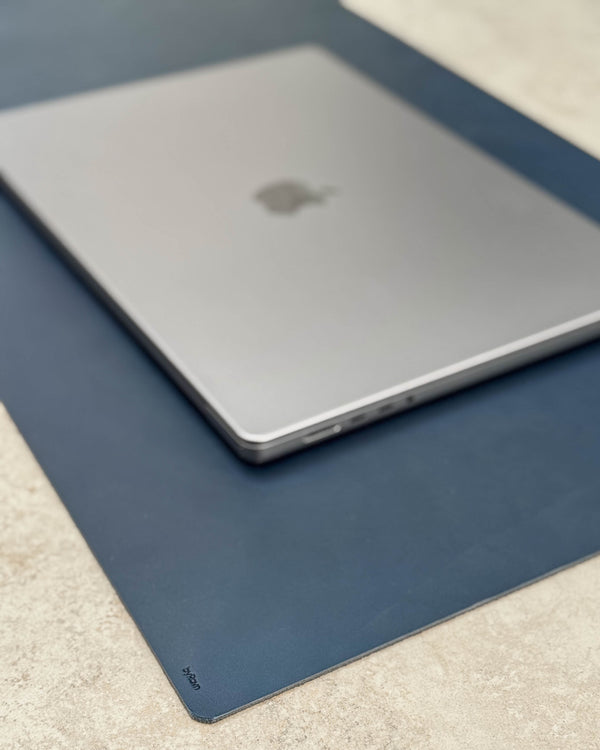 XXL DESK desk pad in navy blue full-grain leather with byRavn embossing under a laptop on a light tabletop