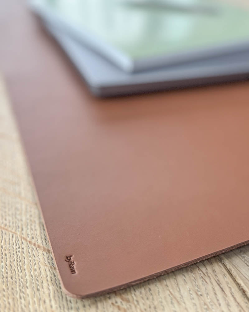 DESK desk pad in cognac full-grain leather, close-up of the corner with byRavn embossing under a laptop on a wooden table
