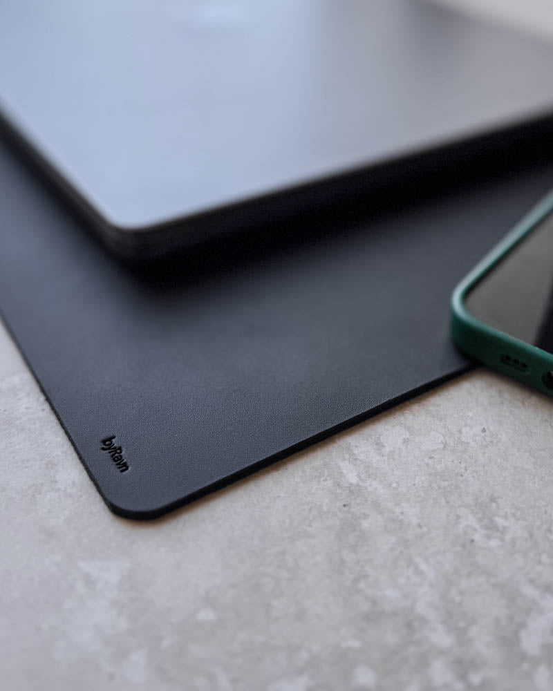 DESK desk pad in black full-grain leather, close-up of the corner with byRavn embossing under a laptop and phone