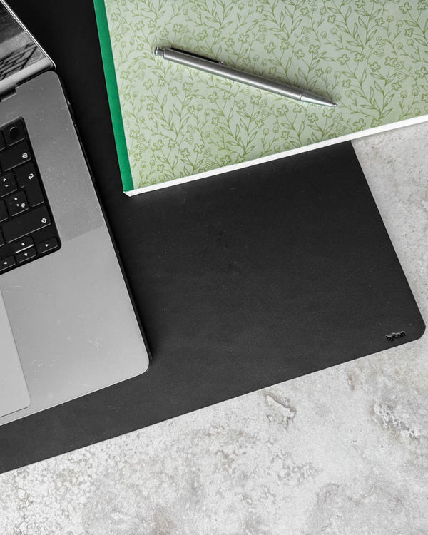 DESK desk pad in black leather seen from above with laptop, floral notebook and ballpoint pen on a light tabletop