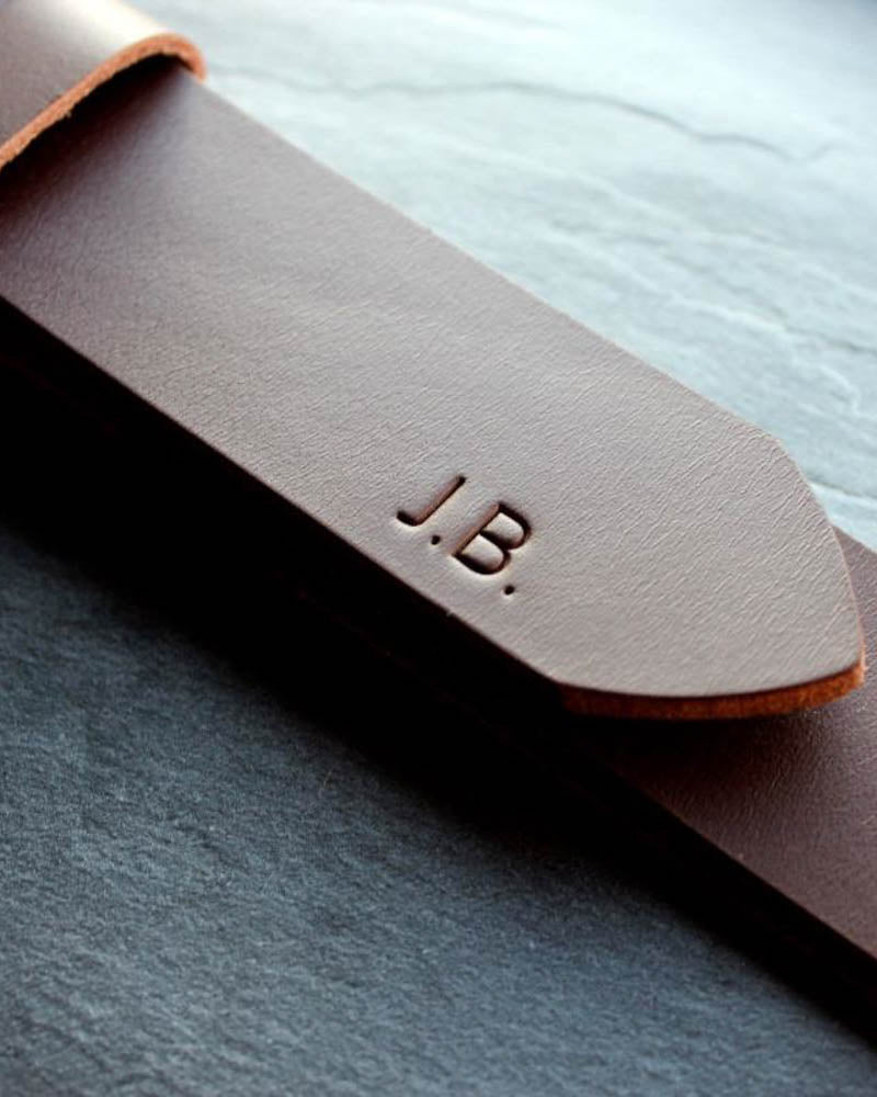Close-up of EDGE belt in dark brown leather with the initials "J.B." engraved on the tip, on a slate slab