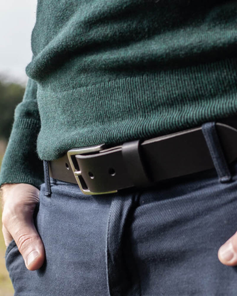 EDGE belt in black leather with steel buckle worn with blue trousers and a dark green knit jumper outdoors