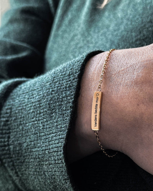 EVI bracelet in gold-plated stainless steel with "Verdens bedste mor" ♡ engraved on a bar plate, worn on the wrist with a green knit