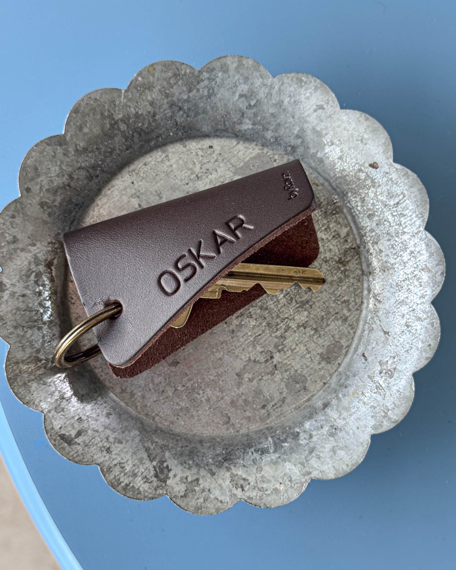 FOLD leather key holder in dark brown with the name "Oskar" engraved, with keys in a zinc dish on a blue table