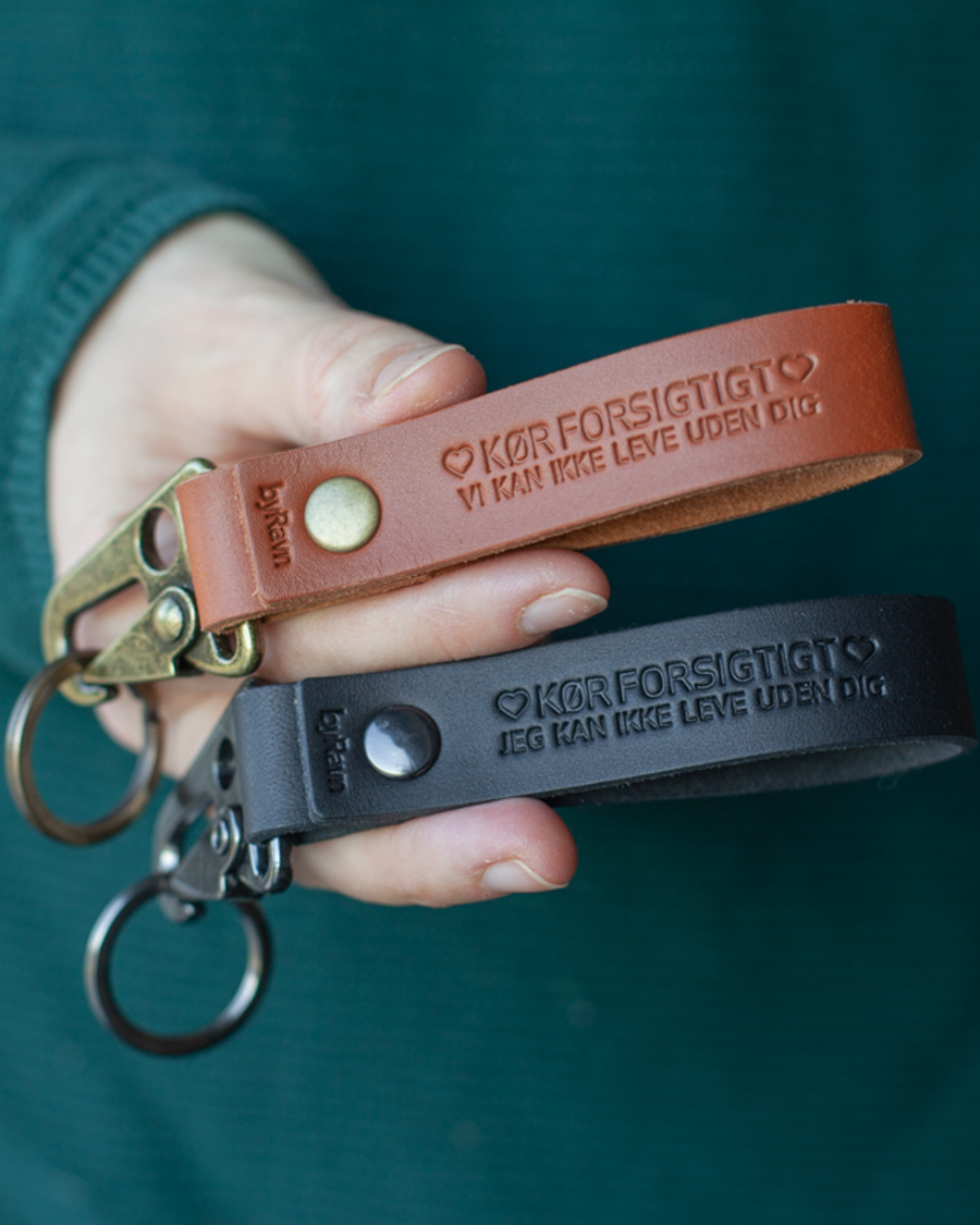 Two HOOK leather keyrings in cognac and black held in hand with the engraving "Kør forsigtigt ♡ vi kan ikke leve uden dig"