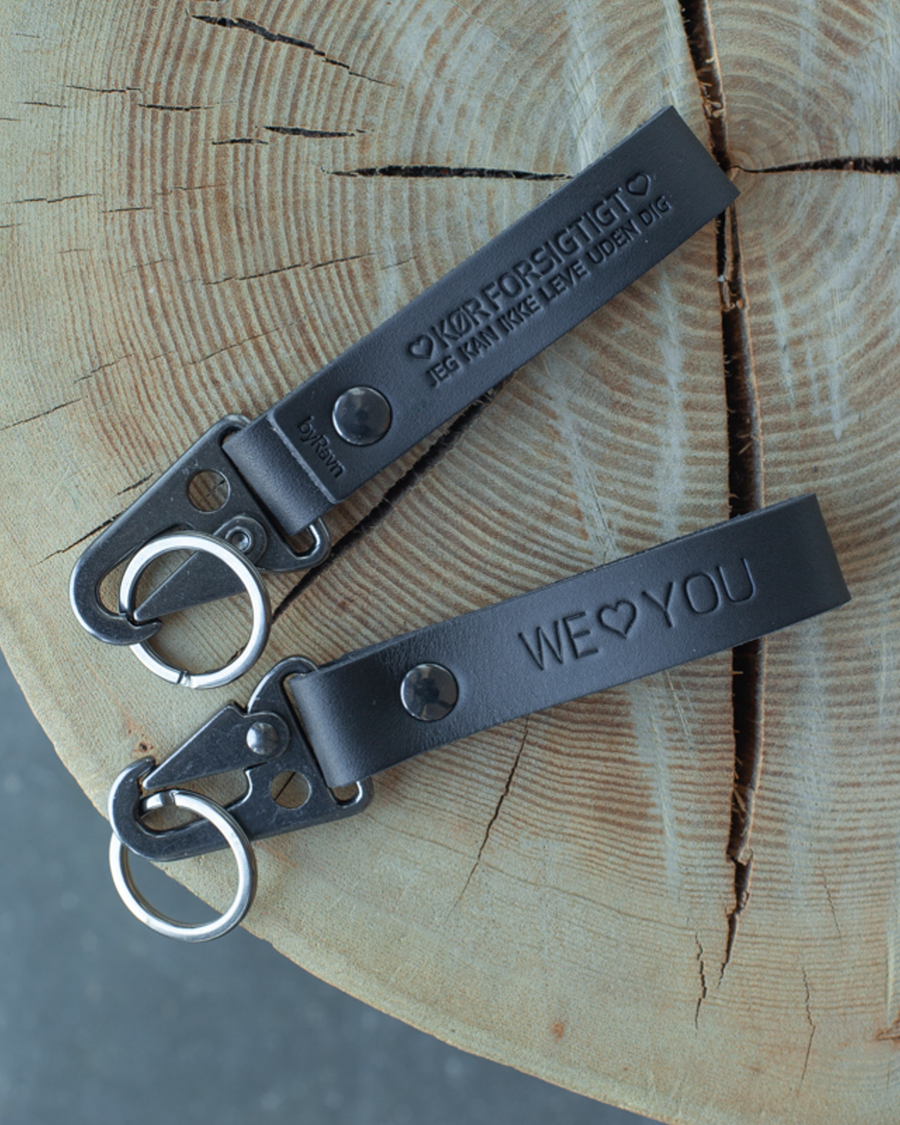 Two HOOK leather keyrings in black on a tree stump with the engravings "We ♡ you" and "Kør forsigtigt – jeg kan ikke leve uden dig"