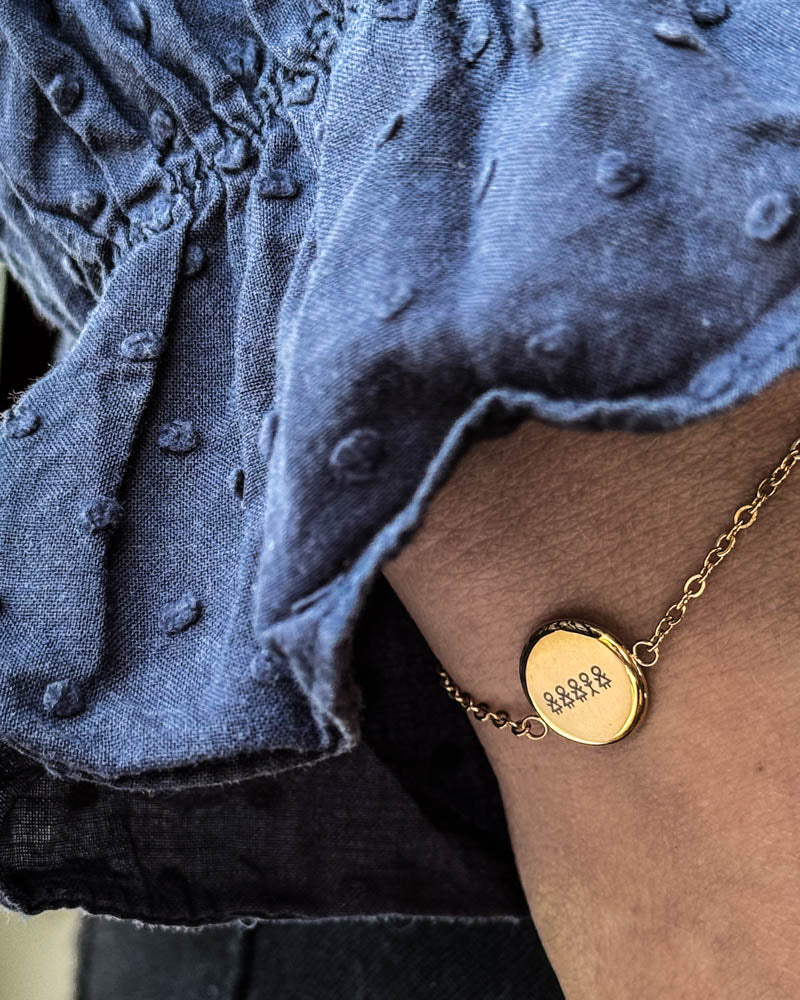 IBI STICK bracelet in gold-plated stainless steel with matchstick family engraved on a round disc, worn on the wrist with a blue blouse