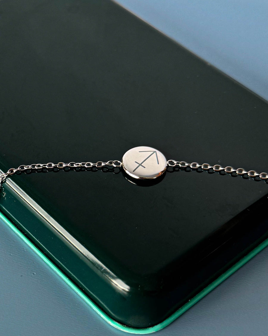 IBI STAR bracelet in silver-coloured stainless steel with the zodiac sign Sagittarius engraved on a round disc, lying on a dark green box