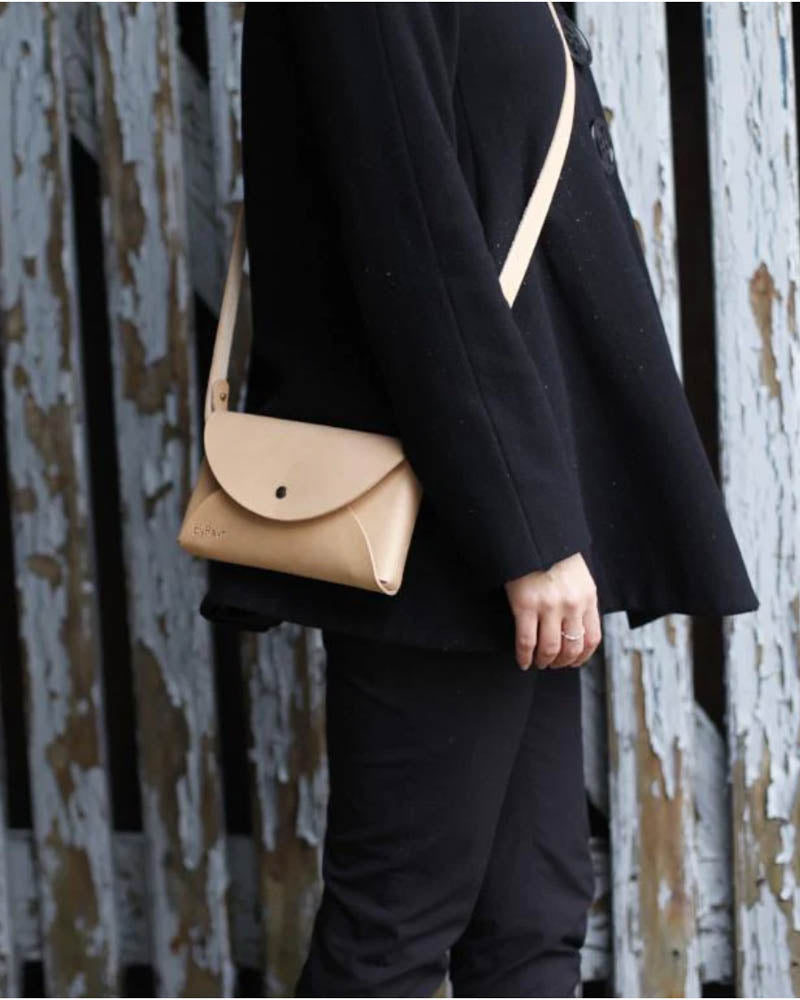 INA shoulder bag in natural leather worn over the shoulder on a black coat in front of a peeling white fence