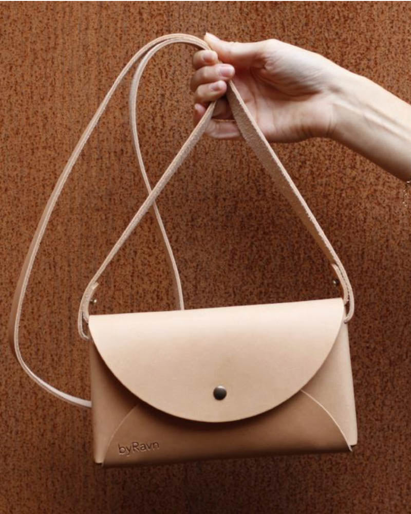 INA shoulder bag in natural leather held up by the strap against a rust-red wall