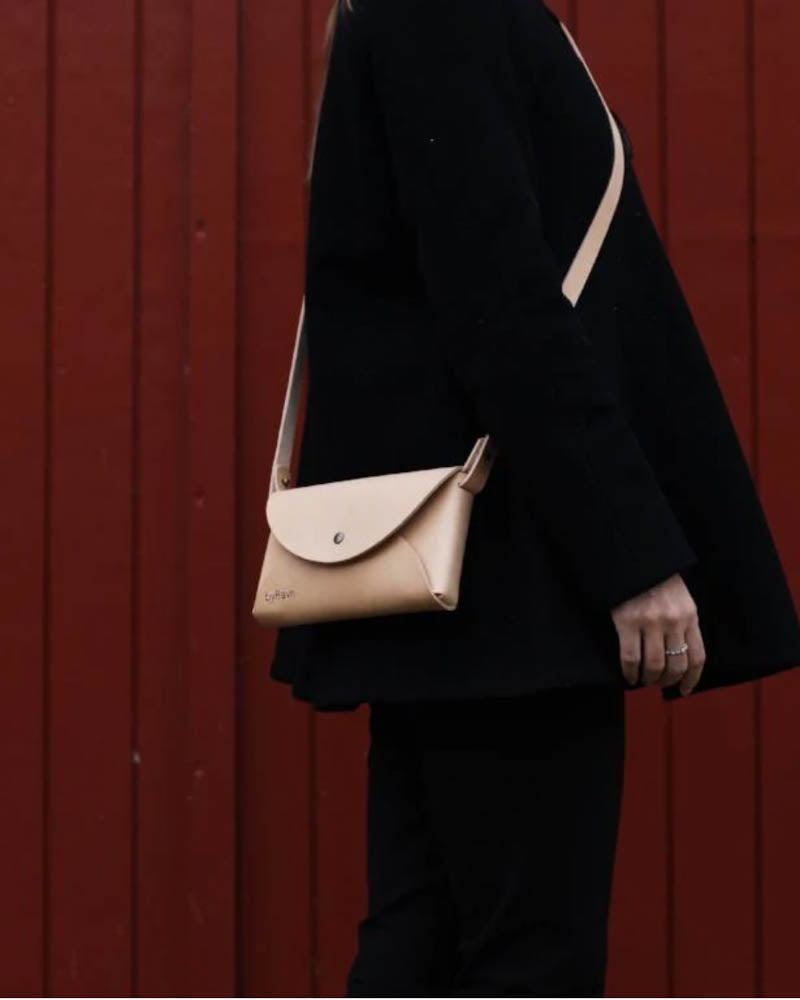INA shoulder bag in natural leather worn crossbody by a woman in black clothes in front of red wood cladding