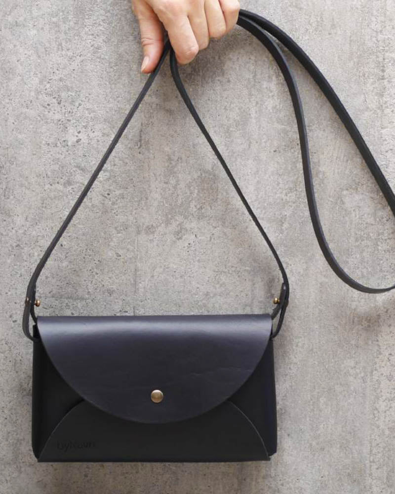 INA shoulder bag in black leather held up by the strap against a grey concrete wall with gold-coloured press stud