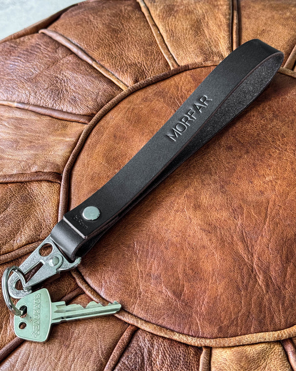 IRON leather keyring in dark brown with the engraving "Morfar" and key, lying on a cognac leather pouf