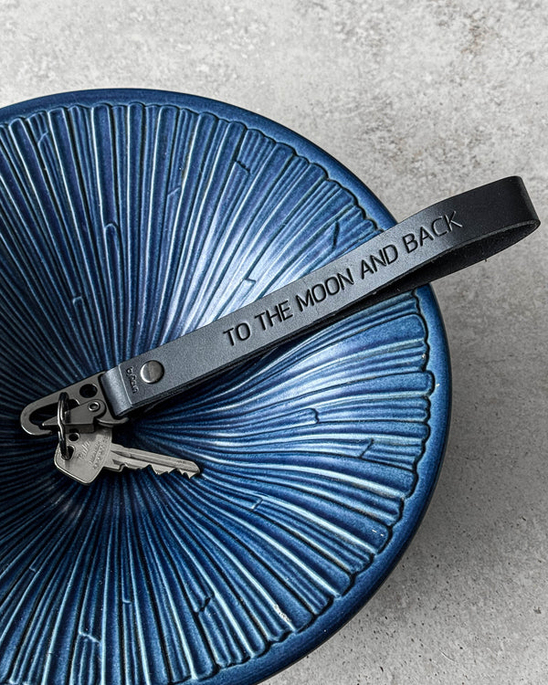 IRON leather keyring in black with the engraving "To the moon and back" and keys in a blue ribbed ceramic bowl