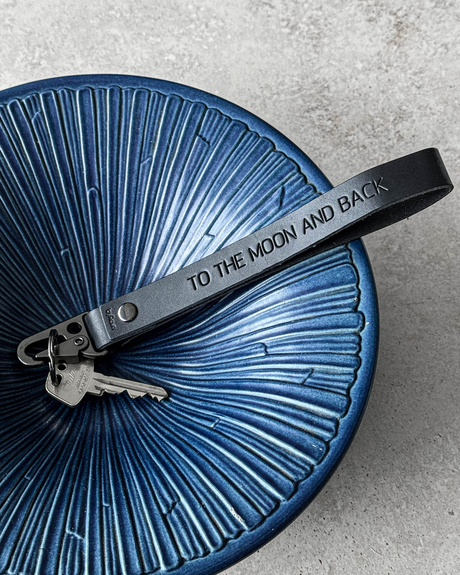 IRON leather keyring in black with the engraving "To the moon and back" and keys in a blue ribbed ceramic bowl