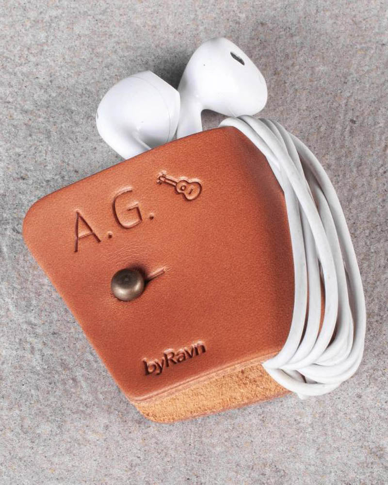 KAX leather earbud holder in cognac with the initials "A.G." and a guitar engraved, with earpods on grey stone