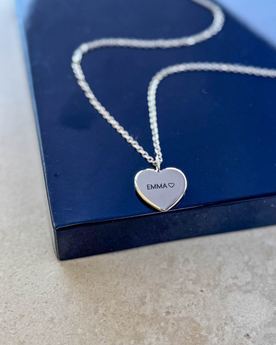 LUV QUOTE name necklace in silver-coloured stainless steel with "EMMA" ♡ engraved on heart pendant, resting on a navy blue gift box