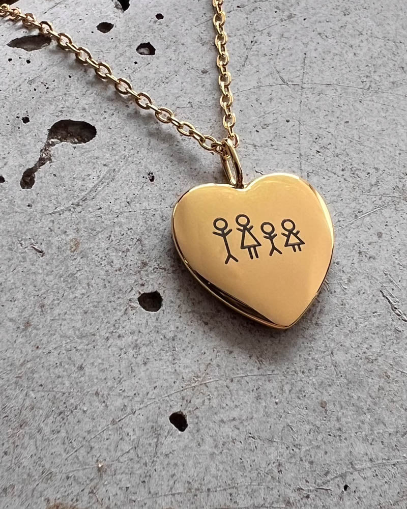 LUV STICK necklace in gold-plated stainless steel, heart pendant engraved with a matchstick family of four, resting on raw concrete