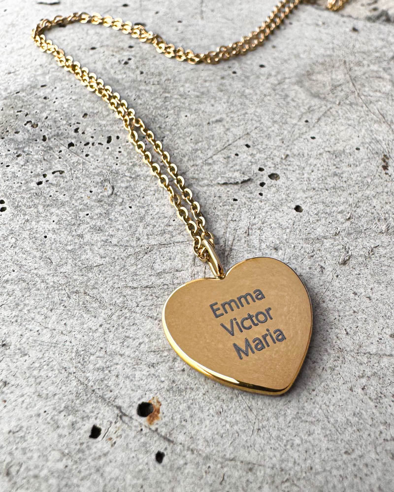 LUV QUOTE name necklace in gold-plated steel with the names "Emma Victor Maria" engraved on a heart pendant, lying on concrete