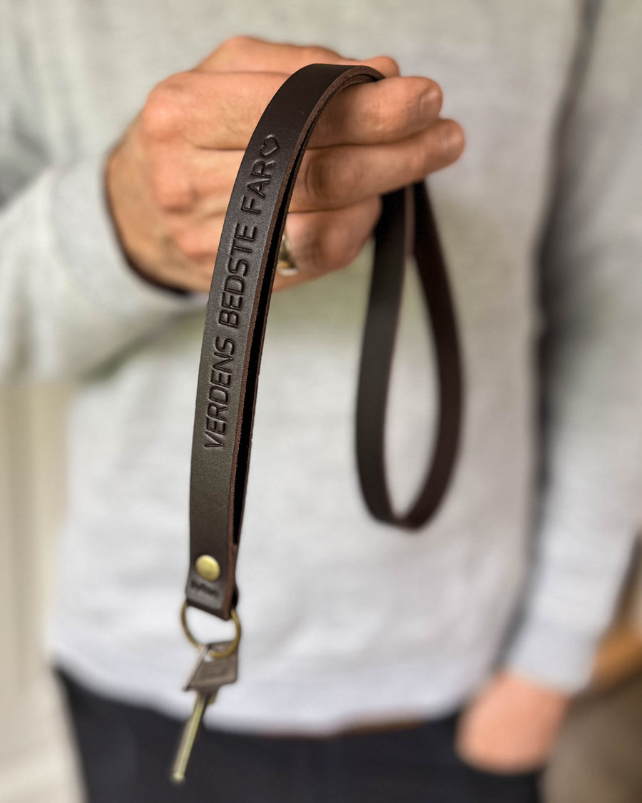 MAX leather keyring in dark brown as a long strap with the engraving "Verdens bedste far" ♡, held in hand with key