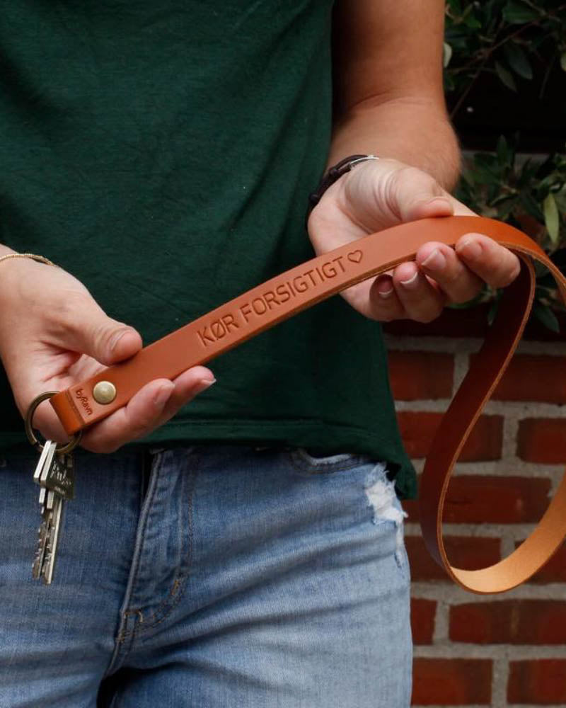 MAX leather keyring in cognac as a long strap with the engraving "Kør forsigtigt" ♡, held in both hands with keys