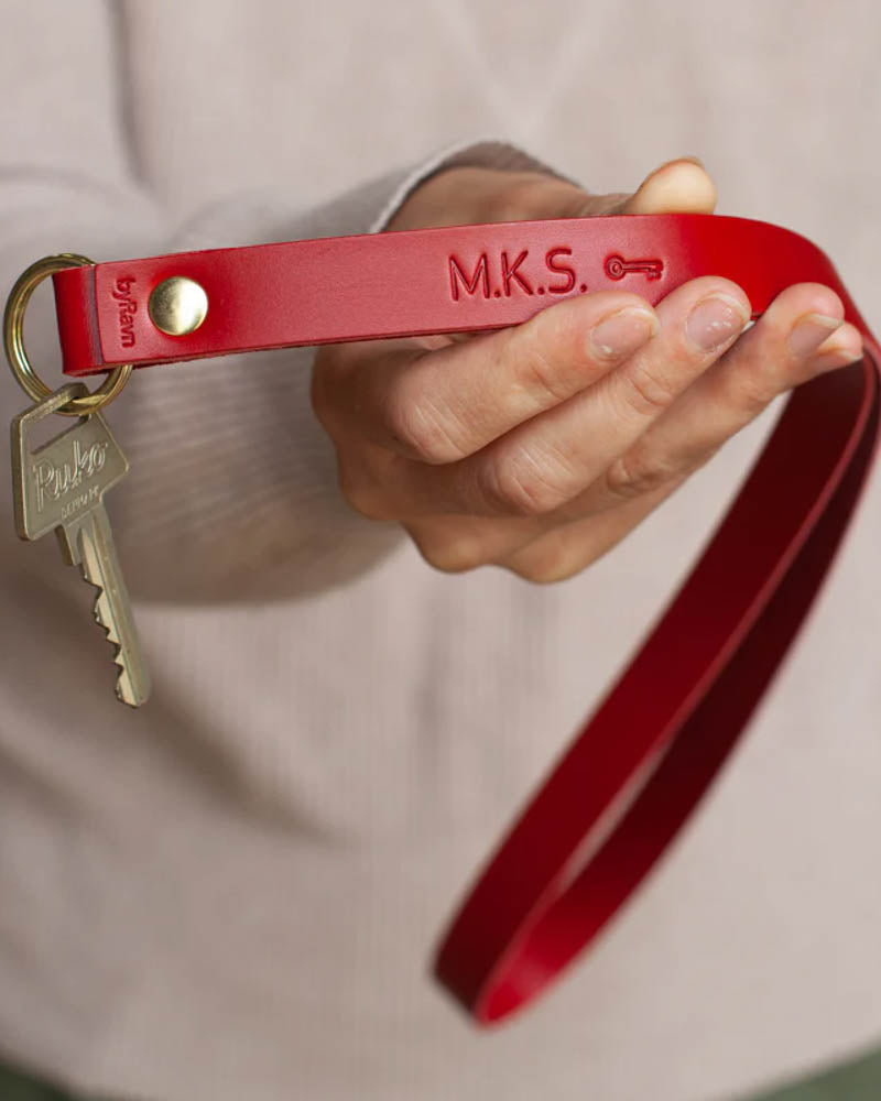 MAX leather keyring in red as a long strap with the initials "M.K.S." and key symbol engraved, held in hand with key