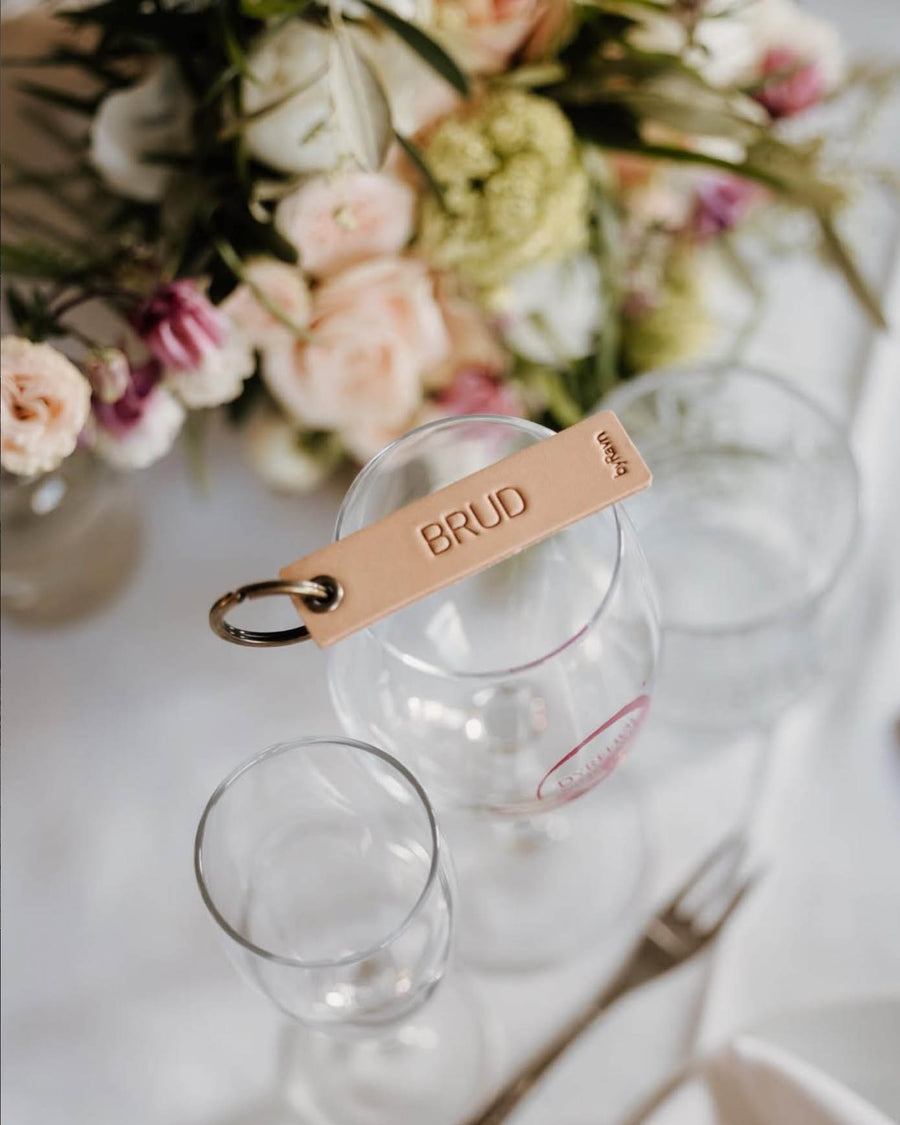MERRY leather place card in natural with the engraving "Brud" and key ring, placed on a wine glass beside a bridal bouquet