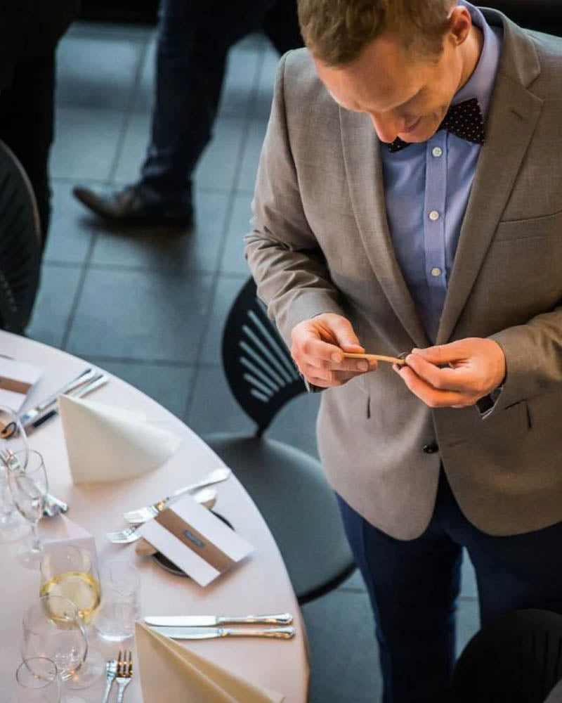 Sample of the MERRY leather place card – guest in suit and bow tie reading his place card at the set party table