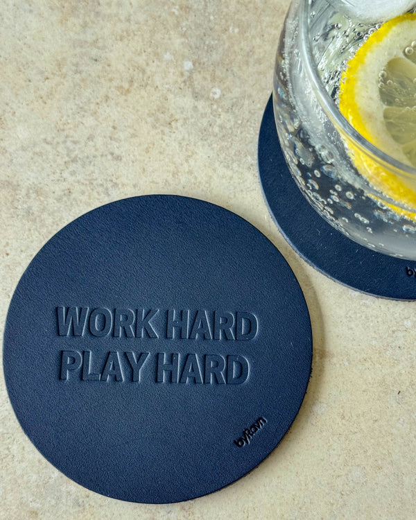 MOKKA coaster in navy blue leather embossed "Work hard play hard" on a light table next to a glass with lemon slice
