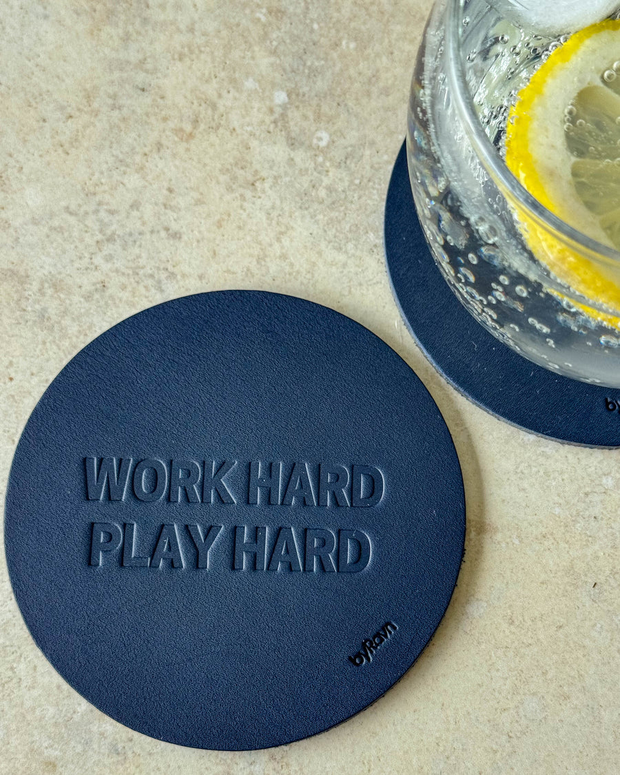 MOKKA coaster in navy blue leather embossed "Work hard play hard" on a light table next to a glass with lemon slice