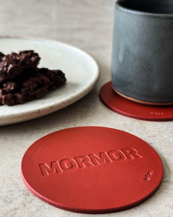 MOKKA coaster in red leather embossed "Mormor" on a light table with a grey coffee cup and a plate of chocolate cake