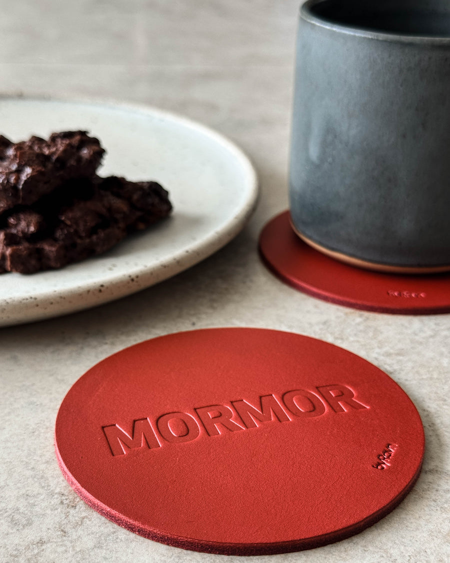 MOKKA coaster in red leather embossed "Mormor" on a light table with a grey coffee cup and a plate of chocolate cake