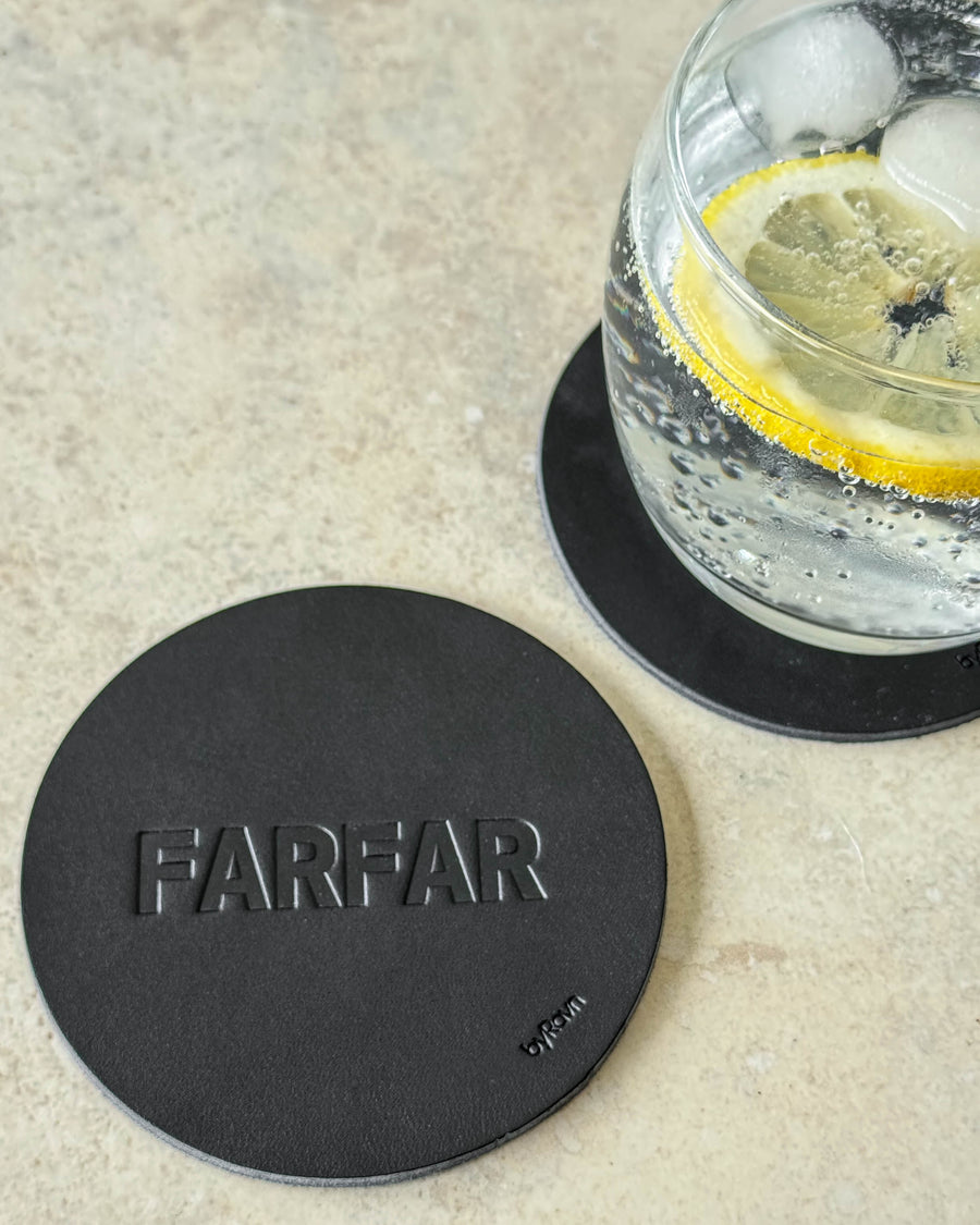 MOKKA coaster in black leather embossed "Farfar" on a light table next to a glass of gin and tonic with lemon slice