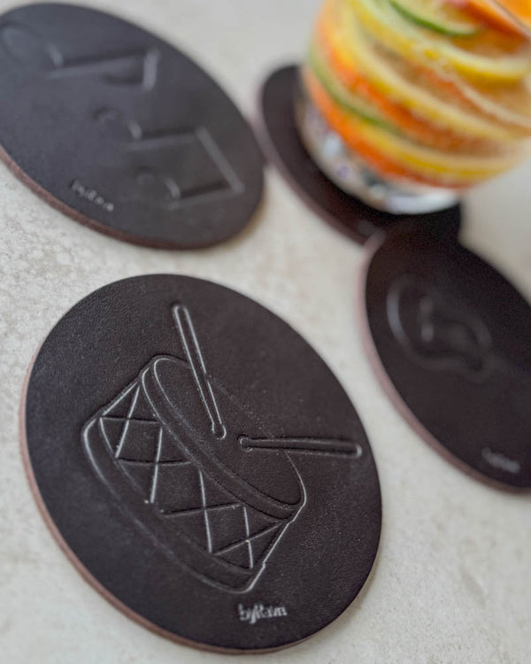 MOKKA music coasters in dark brown leather embossed with drum and sheet music by a glass with citrus slices