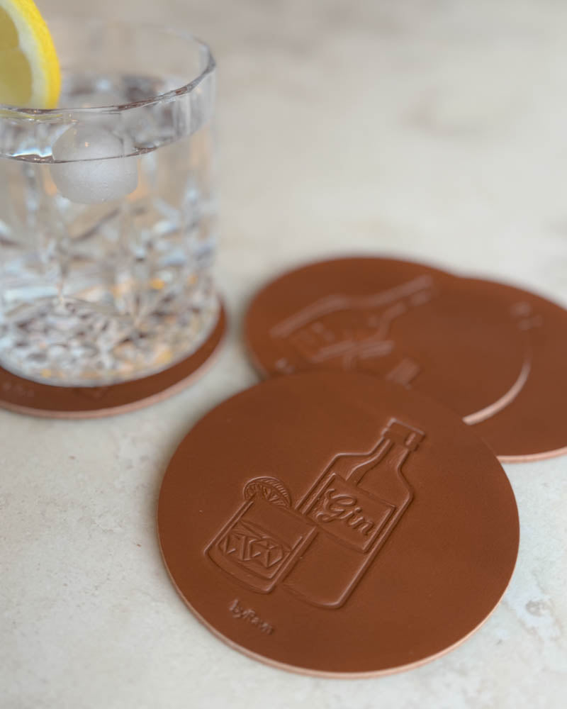 MOKKA spirits coasters in cognac leather embossed with gin bottle and drink, on a light stone surface by a glass with lemon slice