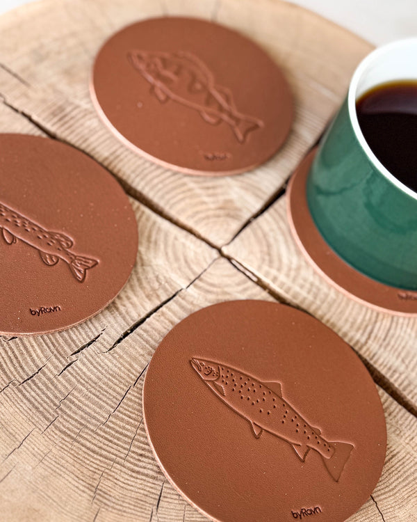 MOKKA angler coasters in cognac leather embossed with sea trout, pike and perch on a wooden slab next to a green coffee cup