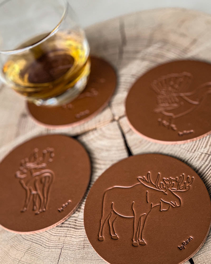 MOKKA hunting coasters in cognac leather embossed with elk, fallow deer and capercaillie on a wooden slab by a glass of whisky