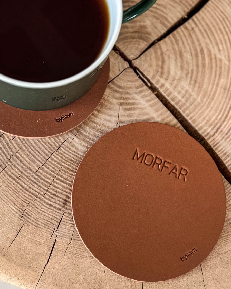 MOKKA round coaster in cognac leather with the engraving "Morfar" on a wooden slab next to a green coffee cup on a coaster