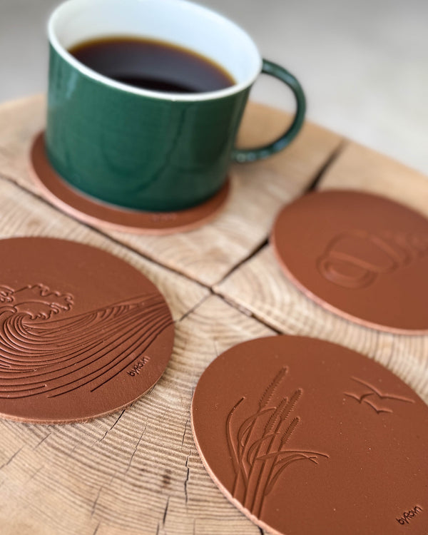 MOKKA summer house coasters in cognac leather embossed with wave, marram grass with seagulls and conch shell on a wooden slab by a green cup