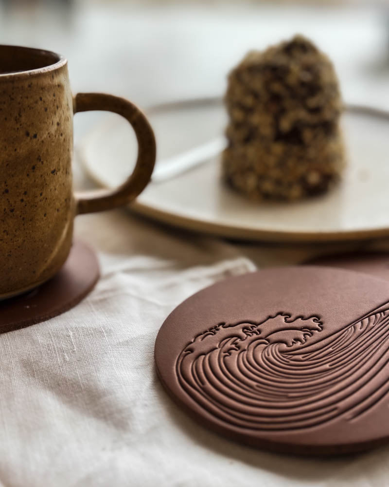 MOKKA summer house coaster in cognac leather embossed with a wave motif on a linen cloth next to coffee cup and plate of cake